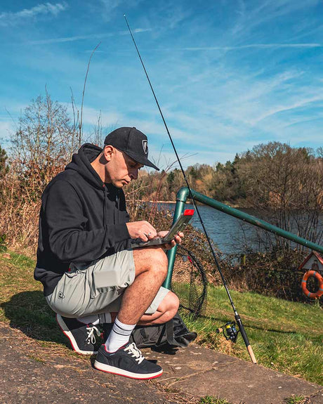 Hombre sentado en un muelle, usando pantalón corto gris, revisando su teléfono junto a una caña de pescar.